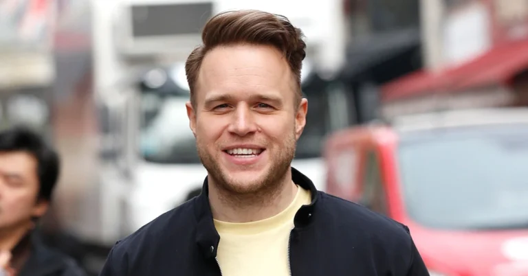 A man with brown hair styled upward, dressed in a dark jacket over a light shirt, is seen walking along a bustling urban street with vehicles and buildings in the background.